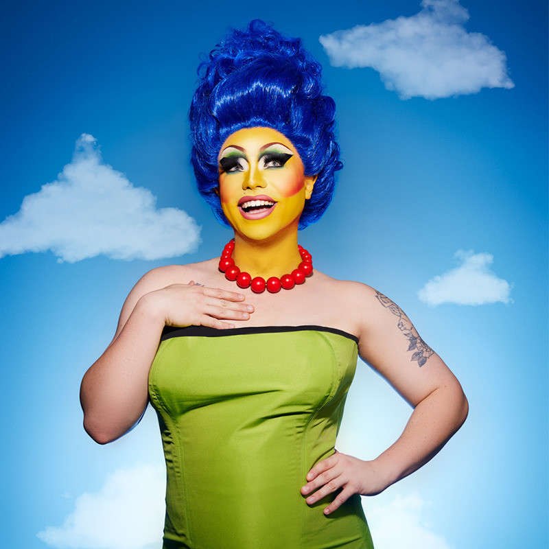 The Stripsons - A drag queen wearing a green dress with orange pearls and large blue hair. Her skin is a bright yellow colour with large exaggerated eye makeup