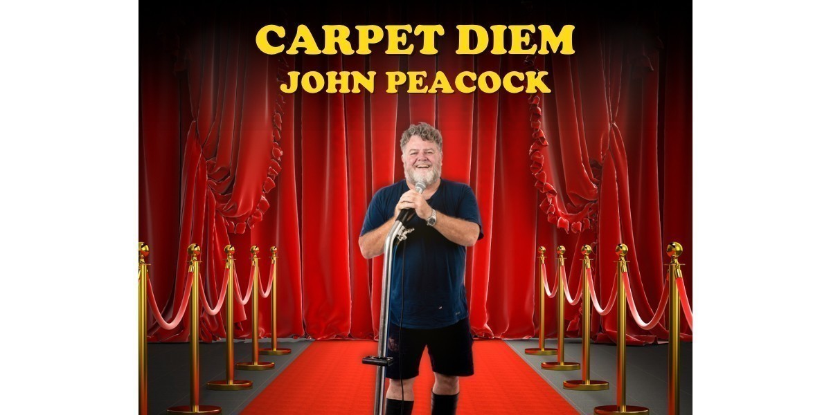 John Peacock stands on a red carpet in front of red curtains holding a carpet cleaning wand.