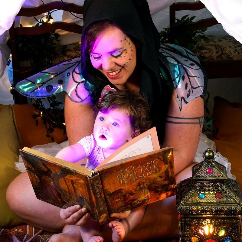 Songs and Stories with Emilie Kate - Emily Kate is sitting cross legged under a white sheet over chairs to make a tent/cubby house. She holds her infant daughter Sunny on her lap. Emily holds a book in front of Sunny for her to read. The book is a Fairy book. Purple light shines onto the baby's face as she looks up in wonder. Emily wears black, blue and green fairy wings and a black hood with sparkly stars on her face. She is looking down at Sunny smiling. Their little tent/cubby house is decorated with fairy lights, mustard coloured cushions and a gold, bejewelled lantern.