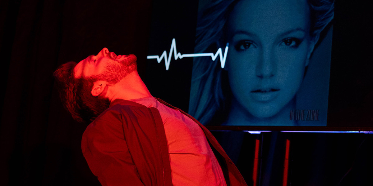 A man leaning backwards kneeling on the floor with a heartbeat monitor and an image of Brittany Spears in the background on a TV screen.