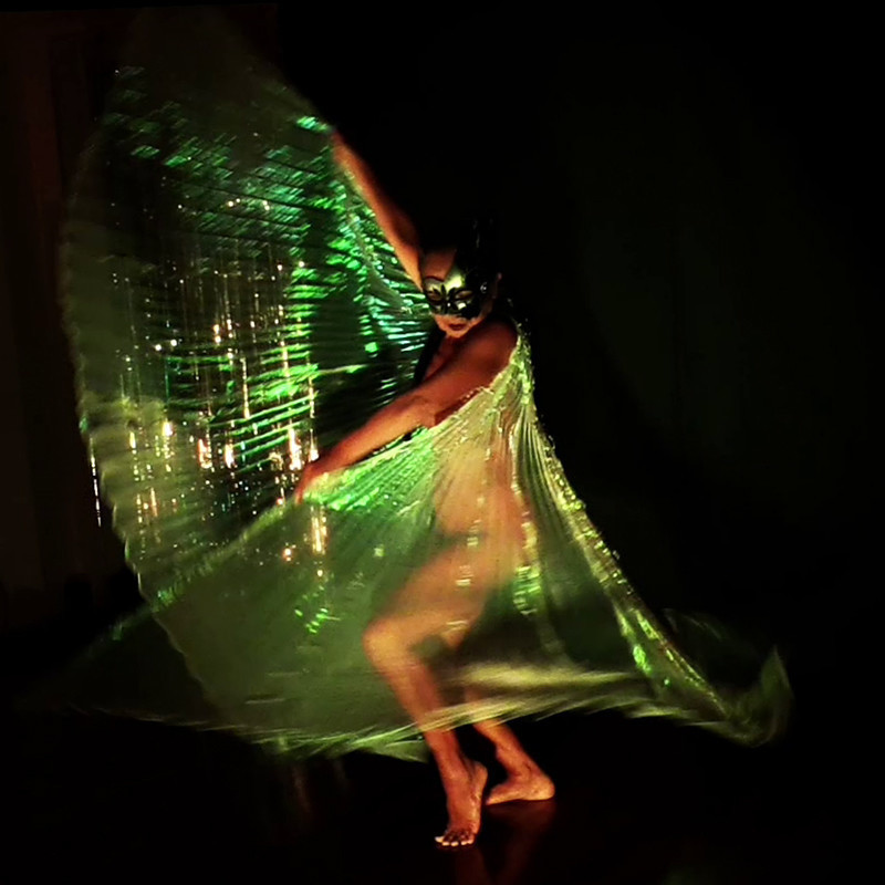 A dancer in a black mask swirling a glowing greenish fabric.