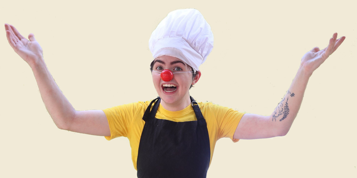A clown faces the camera with both arms raised up to each side in a welcoming pose, with the upper arms laying flat and the forearms on an angle. They have a tattoo of a hare and leaves on their left forearm that is partially visible. Their hands are open with a slight curve to the fingers. They are wearing a yellow short-sleeved shirt, a tall white chef's hat and a black apron, as well as a red nose. They have an excited/welcoming expression with an open mouthed smile - their head is slightly angled to the right. It is a mid shot with an off-white background.