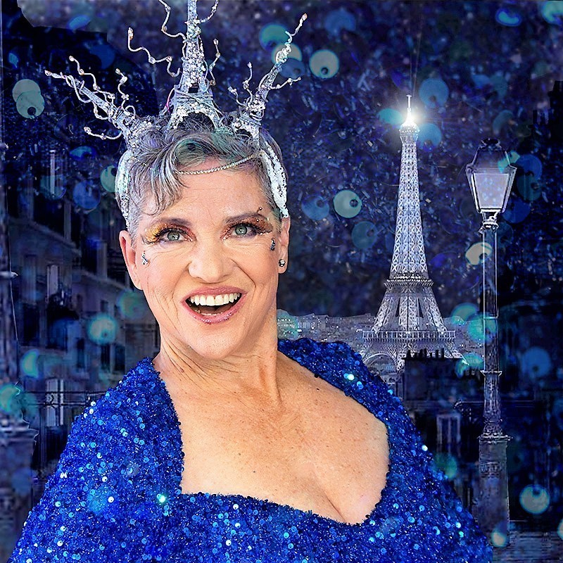 Georgia Darcy, open-mouthed singing, wearing blue-glitter dress and silver-spangled Eiffel Tower tiara.
Eiffel Tower in the background, a Paris sky full of sparkling stars