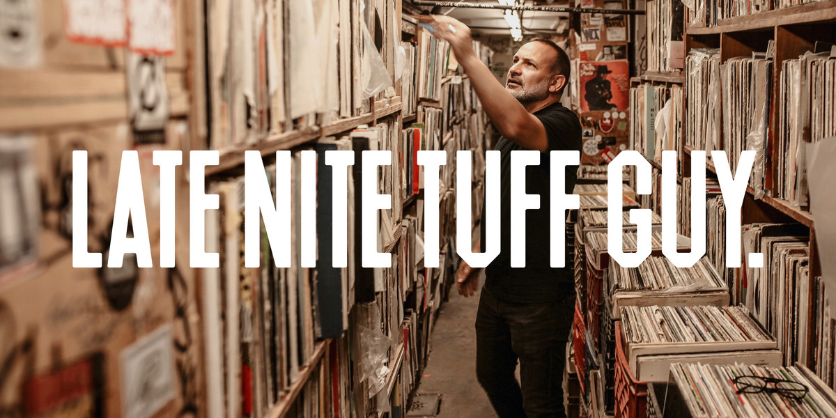 Late Nite Tuff Guy - A man with dark hair and short stubble browses shelves of records, he is wearing black and is looking indistinctly towards the record he's holding