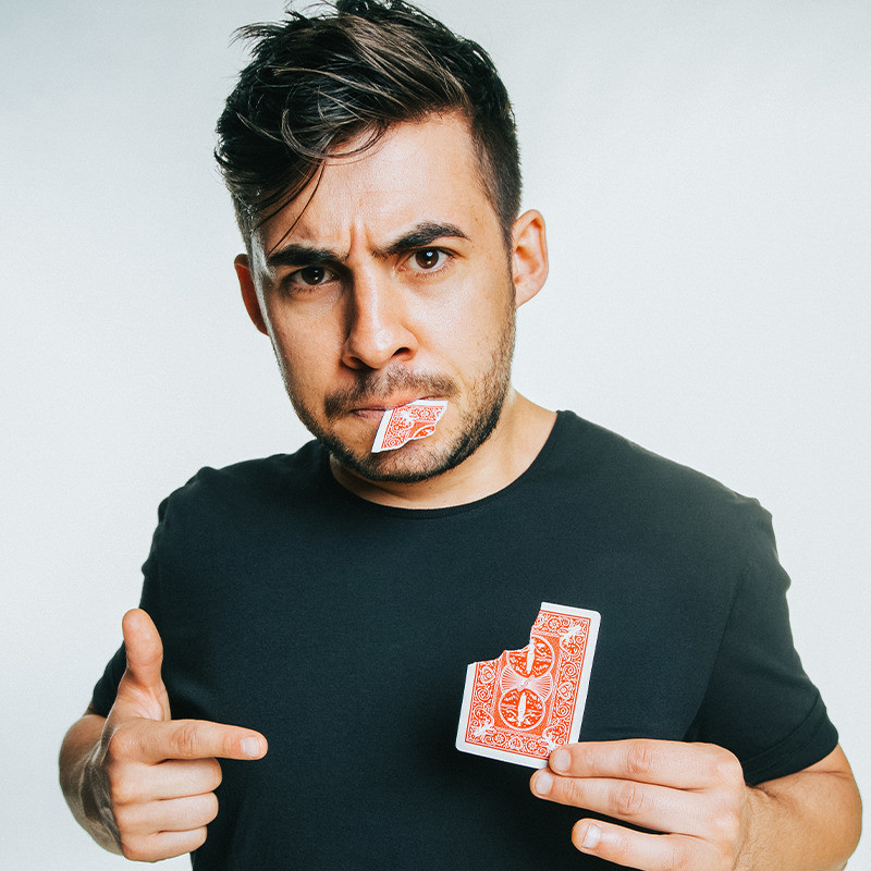 Dom Chambers: A Boy and His Deck - A photo of a person wearing a black t-shirt holding a red playing card in one hand and gesturing to the card with his other hand. The corner of the card is torn off and it’s sticking out their mouth.