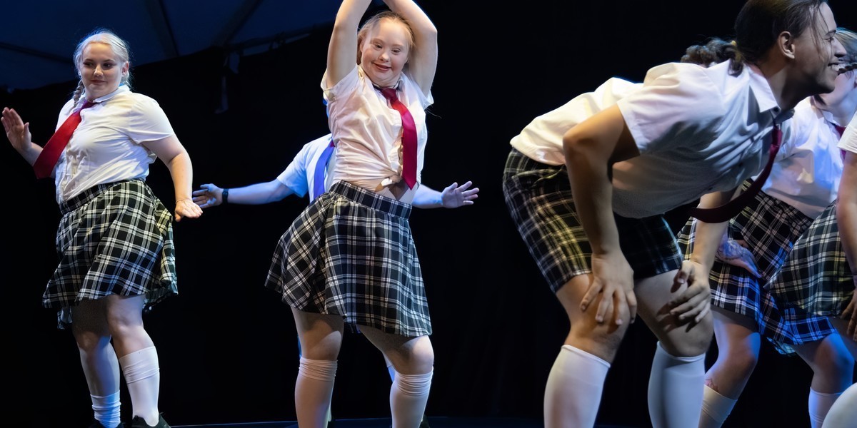 Embrace - A group of artists dancing on the stage, in the center is a young woman in her 20s dancing in a school uniform. Black and white checked skirt with white blouse and red tie. She is surrounded by 3 other individuals and she is smiling.