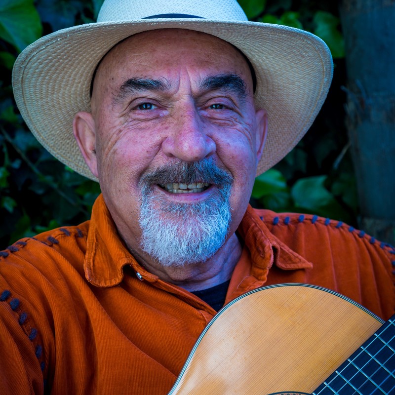 A photograph of a man smiling. He is wearing a cream wide brimmed hat and an orange shirt. He has a grey beard and freckles across his nose. In the right corner there is a glimpse of an instrument.