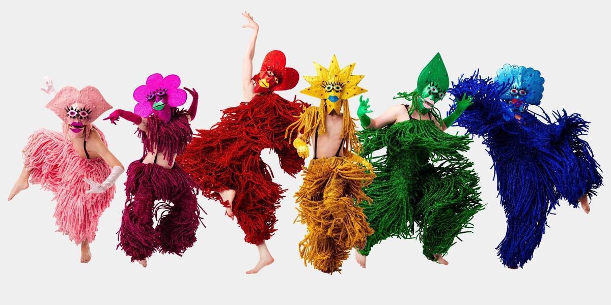 6 performers in a line striking different poses, all in different individual coloured costumes. From left to right: Baby pink performer has a mask in the shape of lips, magenta performer has one in the shape of a flower, red in the shape of a heart, yellow as the sun, green as a leaf, and blue as a cloud.