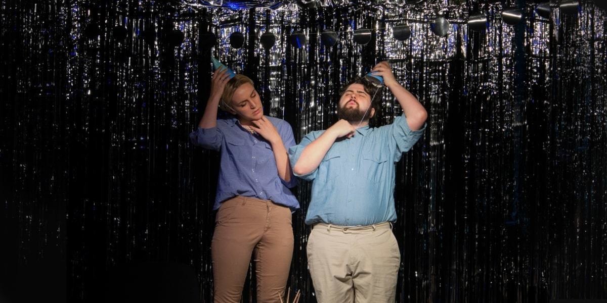 A man and a woman wearing birthday party hats, denim blue shirts and brown pants, standing in front of a sparkly background. They are both taking off their hats.