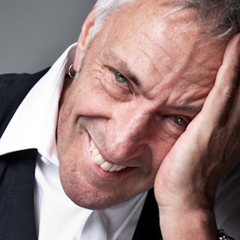 Actor John Waters cradles his smiling face in his left hand. His grey green eyes twinkle mischievously. His face has a bit of stubble and just the right amount of wrinkles to make him ruggedly handsome and sexy. He's wearing a large collared, crisp white shit and a black vest, and has an earring in his right ear.