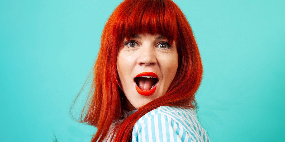 Bron Lewis - 'Obviously' - Bron Lewis with bright red hair and lipstick, stands gasping in front of a turquoise background.