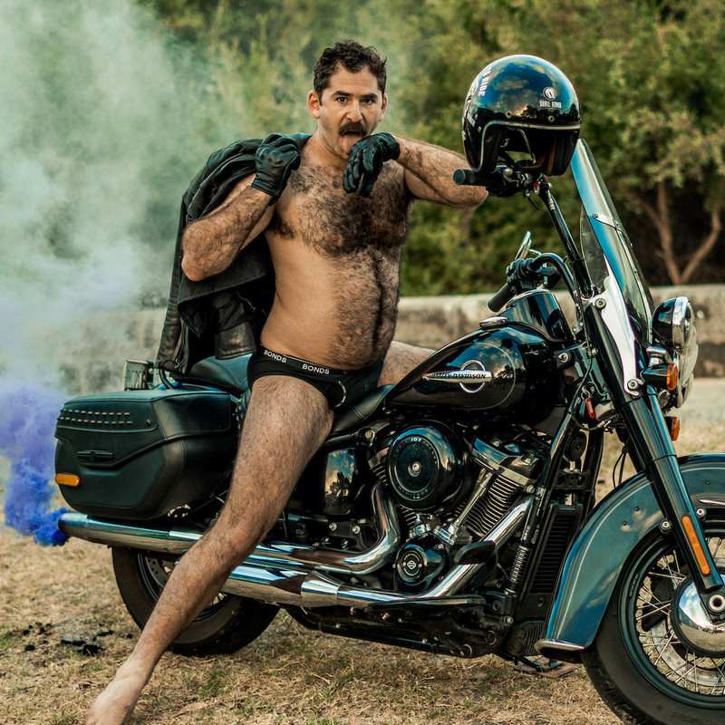 A mostly naked man with black underwear and black gloves on sits on a dark green motorbike, holding a leather jacket over one shoulder, and licking the other hands wrist. Blue smoke is coming out of the exhaust pipe. The man is dark haired and has a hairy chest and arms.