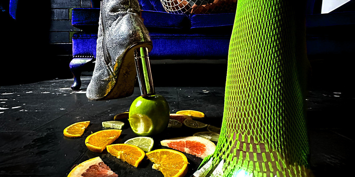 a person in green tights and silver heels steps on a bitten apple with fruit scattered on the ground with a purple couch behind them