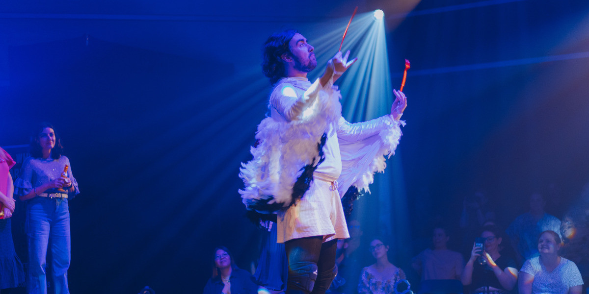 A man stands on stage holding an orange knife and fork. He is wearing wings with white and black feathers.