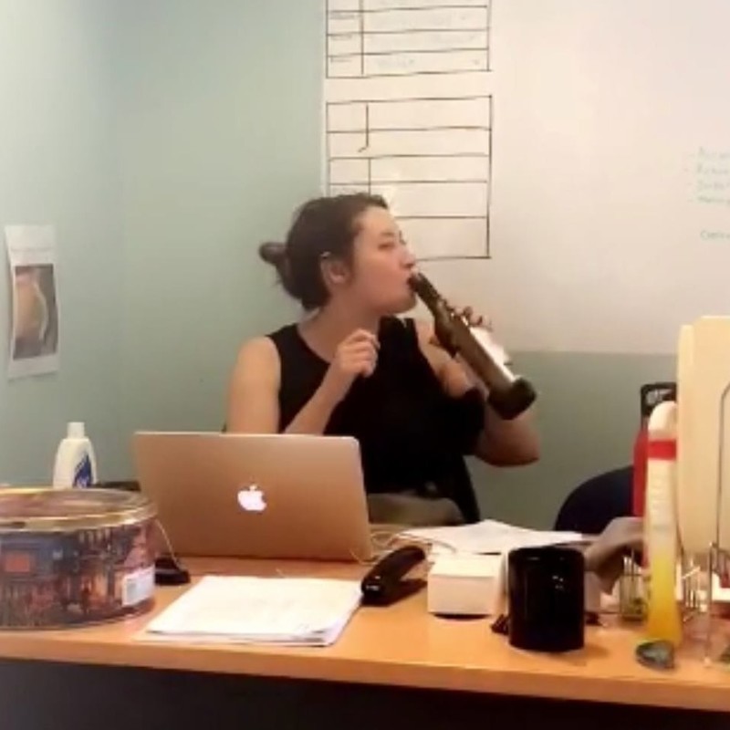 I Thought I Was Having An Existential Crisis, But It Was Just A Gluten Intolerance. - A candid style photo of a woman sitting at a desk in front of a laptop drinking straight out of a wine bottle.