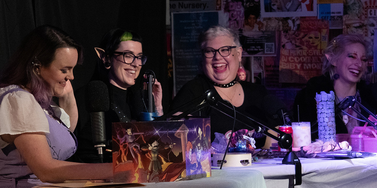 Big Crit Energy - live Dungeons and Dragons! - Four femme people sitting at a dungeons and dragons gaming table looking at each other and laughing.