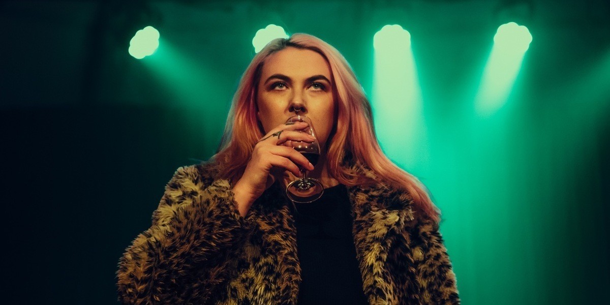 Fiona stands on stage wearing a fur coat, looking angry and holding a glass of wine