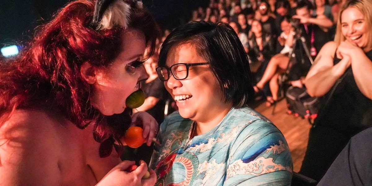 A close up photograph of two people. The person on the left has red hair and is dressed up as a feline with a round shaped item in their mouth. The person on the right is laughing and has short black hair, black framed glasses and a blue top. In the background there is an audience of people.