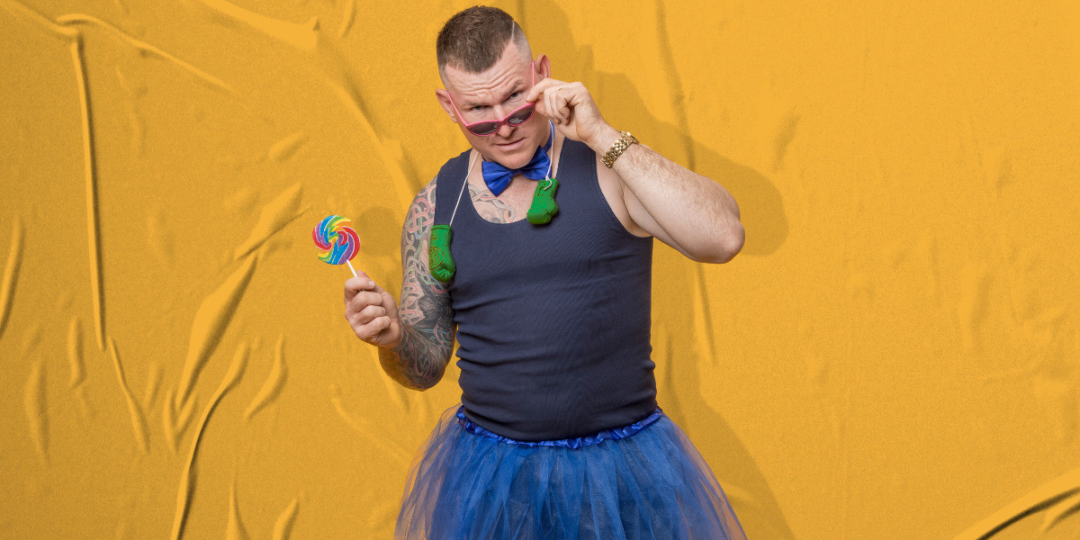 Did You Just Assume My Personality? - one man standing central, wearing a blue singlet, blue tutu, blue satin bow tie, mini green boxing gloves hanging around his neck, holding a round large lollypop with his right hand out to the right of his head, and pulling a set of pink sunglasses down onto the bridge of his nose looking into the camera with an expression as to say "did you just say what I think you did" on his face. 
A yellow crinkled back drop as the background.