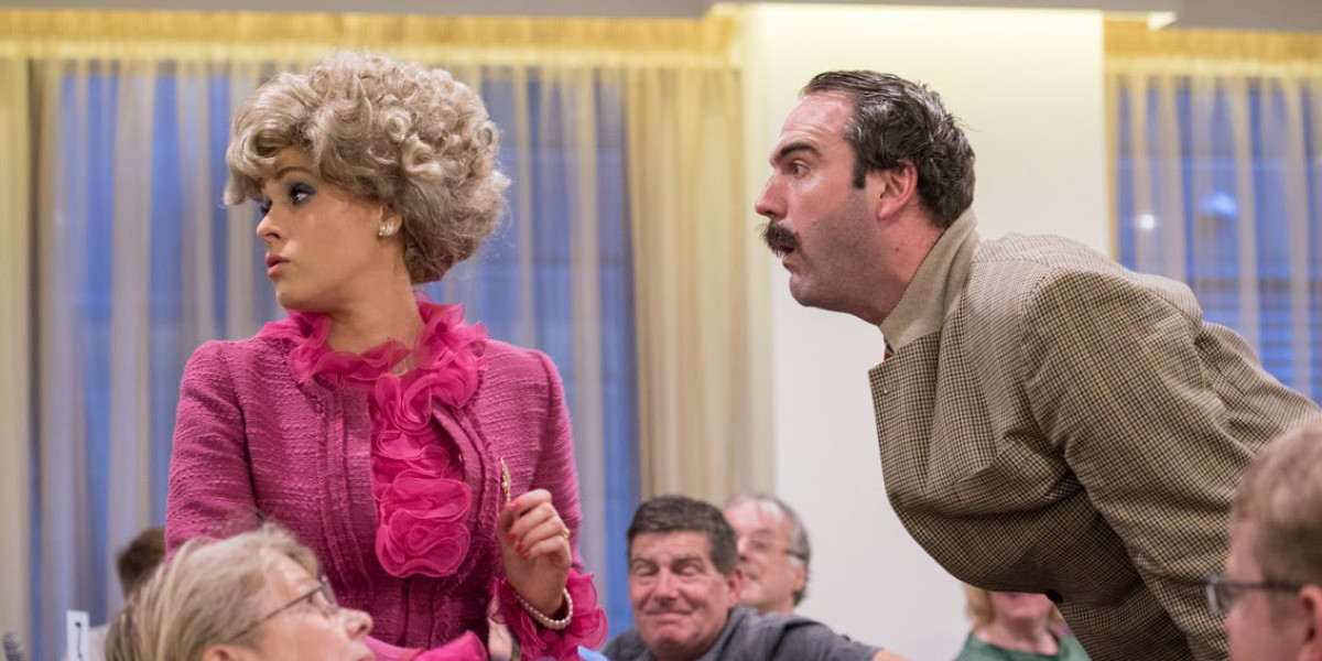 Sybil wearing a pink frilly top looks away from Basil who is screaming at her. Audience can be seen seated around them smiling on at the scene.
