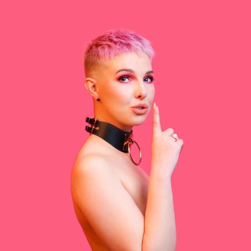 Lauren Thiel: Don't Tell Dad - Lauren stands against a hot pink background, holding her hand to her mouth in a 'shhh' action, implied nude, with a bdsm collar.
