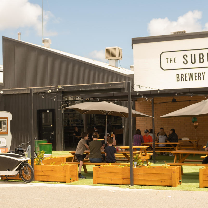 This image is of people sitting outside under a pergola. There is a sign for the brewery at the top of the building and people drinking some beer in the sunshine.