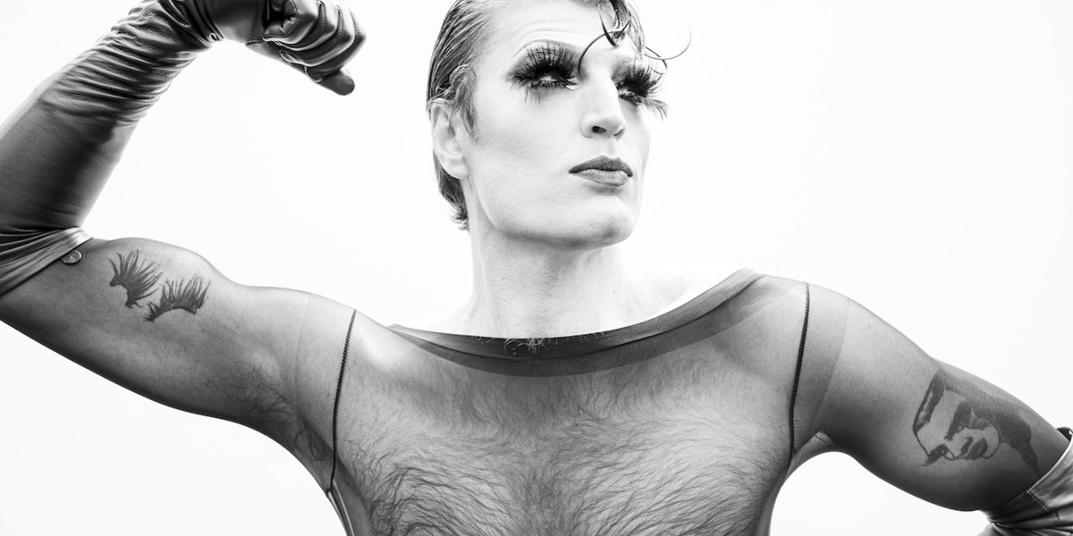 Man with drag make up on and false eyelashes stands looking to the left with his Right arm held up. You can see tattoos on his arms under a black mesh top. Black and white image
