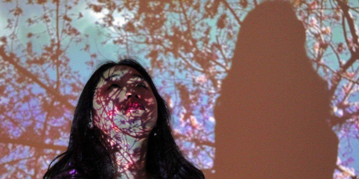 An Asian woman stands just off centre of the image with her gaze facing upwards. She has black hair and has a curious expression on her face. Her shadow is reflected on the right side of the image. An image of a cluster of branches against a blue, pink and and purple sky is seen projected onto the face and neck of the woman, as well as her background.