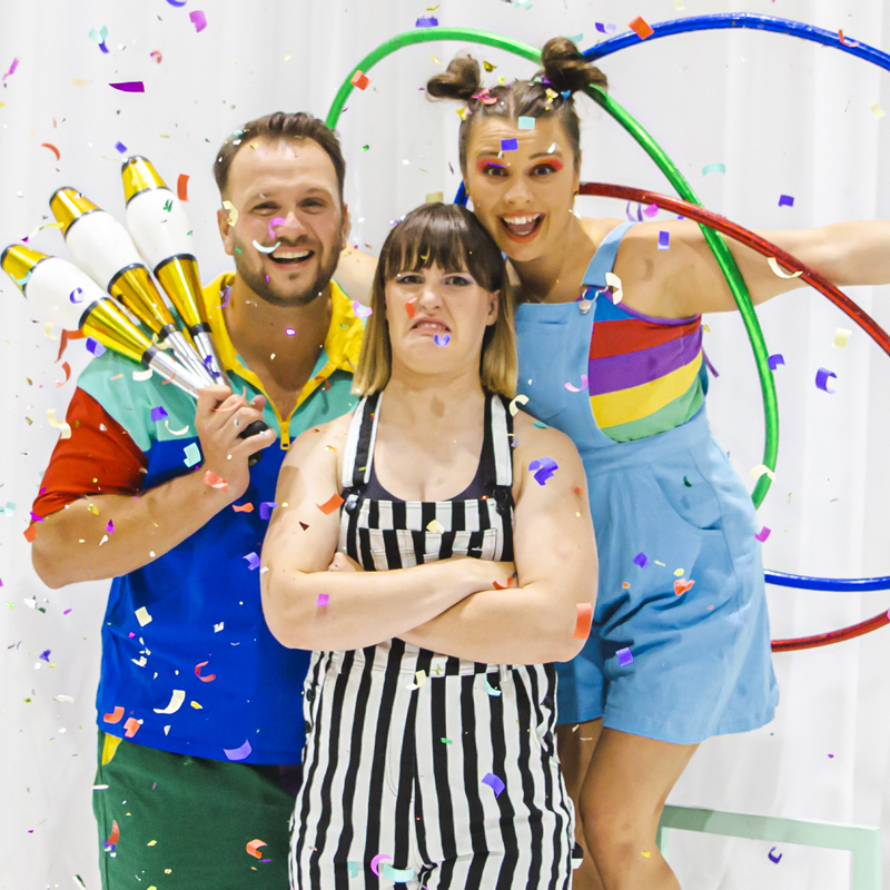 3 people stand looking down the camera, two are smiling wearing colourful clothes. The middle is frowning and glaring in black and white. Confetti falls all around and in front of them.