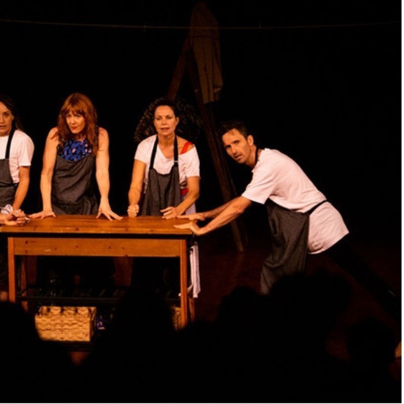 A Picture of 5 people around a wooden table on stage. On the left, a man hunched over with his hand on the table, a woman to his right in a white shirt with a black apron, and to her left a woman with red hair wearing a black apron with a blue floral shirt underneath. A woman on the right with a white shirt and black apron with a man to her right-leaning on the table also in a white shirt and black apron.