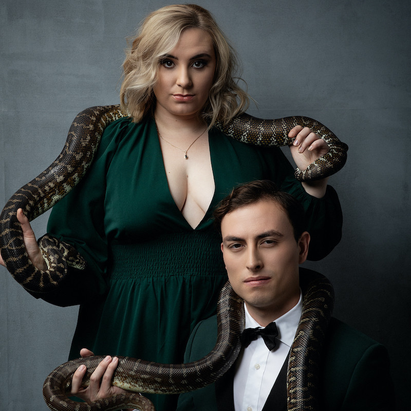 The cast of Snake Pit is posed in front of a grey background. Maddie Houlbrook-Walk, a blonde-haired woman is standing on the left wearing a flowing green dress with a live snake draped on her shoulders. John Robles, a brown-haired man, is seated in front of her in a green tuxedo also with a live snake around his shoulders. They both look towards the camera with serious expressions.