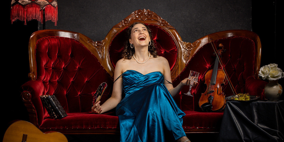Sophie, a young white woman with brown hair is sitting on an ornate red velvet couch. She is wearing a silky blue gown with her legs crossed, one hand holding a floral hand fan and the other a wine glass. She is looking up with her mouth open as if she is mid laugh. On the left there is a classical guitar on its side and an old red fringe lamp shade on a lamp stand, and on the right, her black shoes on the floor and a black tablecloth covering a stool. On top of the stool are some grapes, a wine glass and some red roses in a vase.