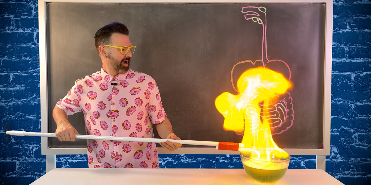 A bearded pan wearing a pink shirt with a doughnut print is standing in front of a chalkboard. He is holding a candle to a bowl of bubbles, which have burst into flames.