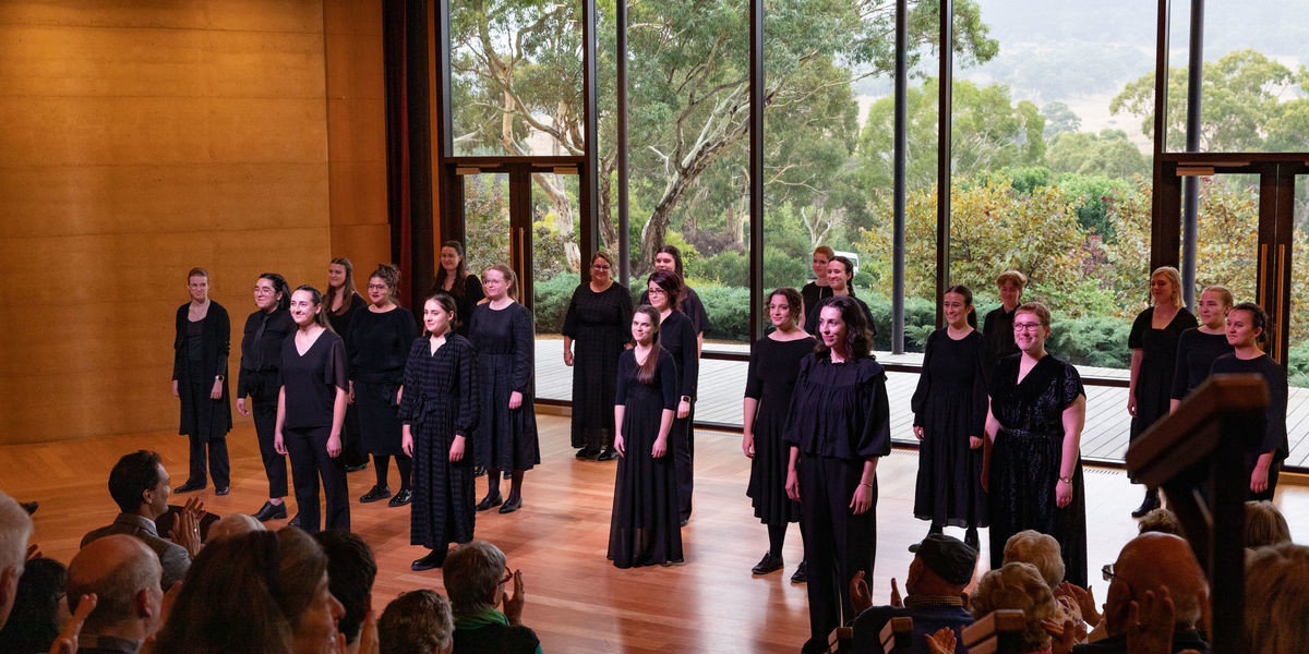 Aurora Vocal Ensemble smiling after performing at Ukaria Cultural Centre. They are standing staggered across the space, with a view of the Adelaide Hills in behind them. They are wearing formal black outfits.