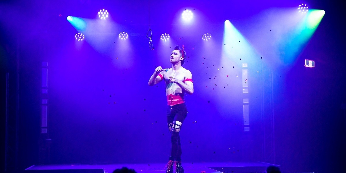 A performer in high heels approaches an aerial appartus. The lighting is dim blue and there is confetti in the air.