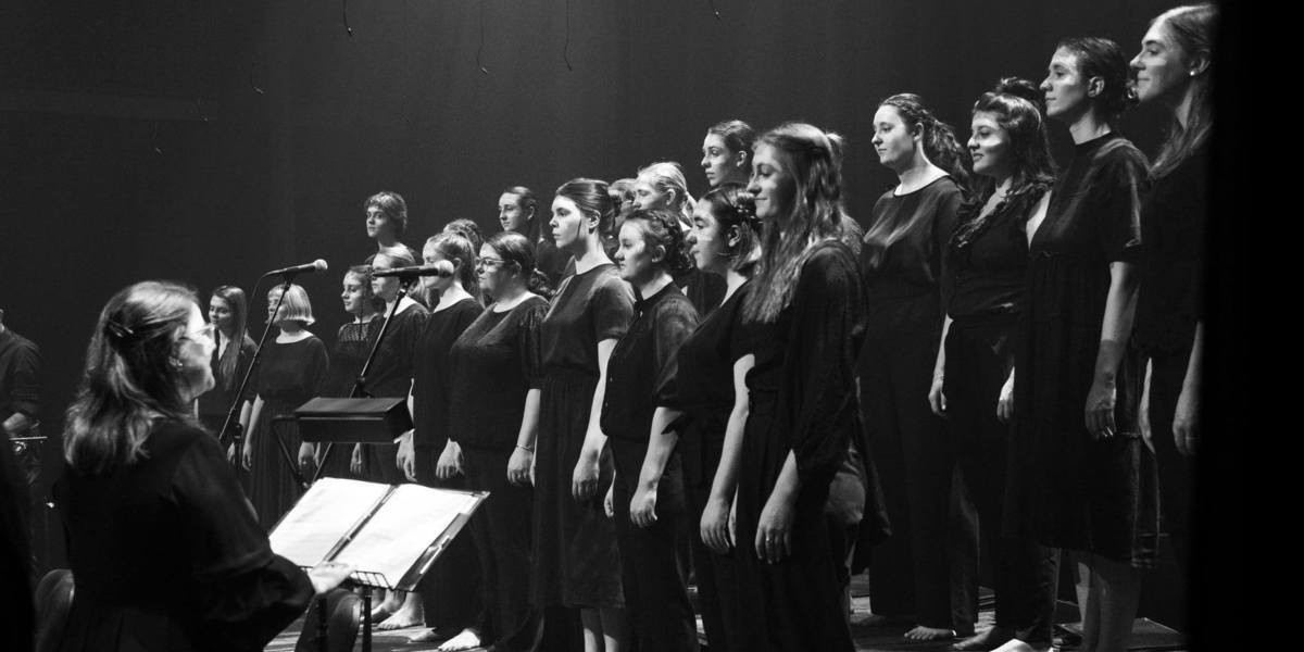 A black and white photo of Aurora Vocal Ensemble and their conductor Christie Anderson. They're standing on a stage with neutral expressions. The photo is taken from the side of stage, with the profiles of the groups faces in focus. They have traditional paint on the foreheads and noses, painted by Galpu elders and family.