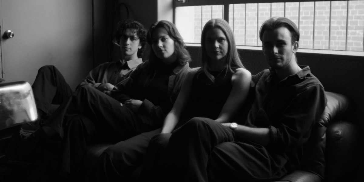 Black and white image of band members sitting side by side on a couch, soft light coming through the window.