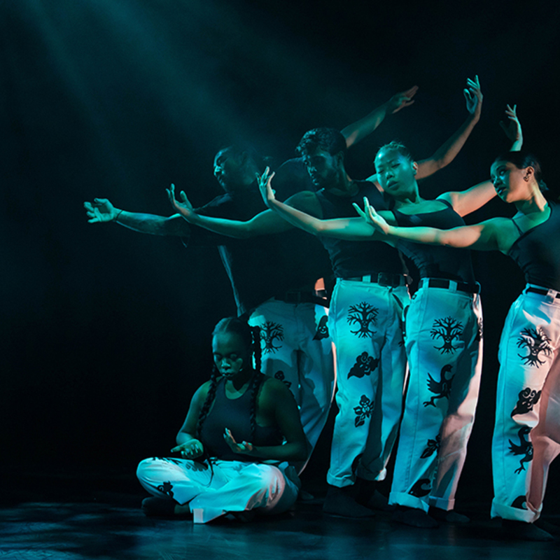 Four dancers stand in a staggered line, their arms are up in the air like a wave. They stand over another woman who is sitting on the floor.