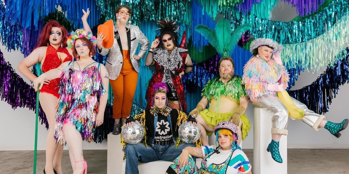 A group of artists pose very seriously as if they were on the cover of Vogue together dressed in colourful sparkly clothing in front of a streamer installation of shimmery greens and blues. Artists include from left to right Cleo Taurus, Diana Divine, Arthur Nicely, Scott Balls, Say Gah, Calamity Tash, Mathew Barker and Annie Schofield