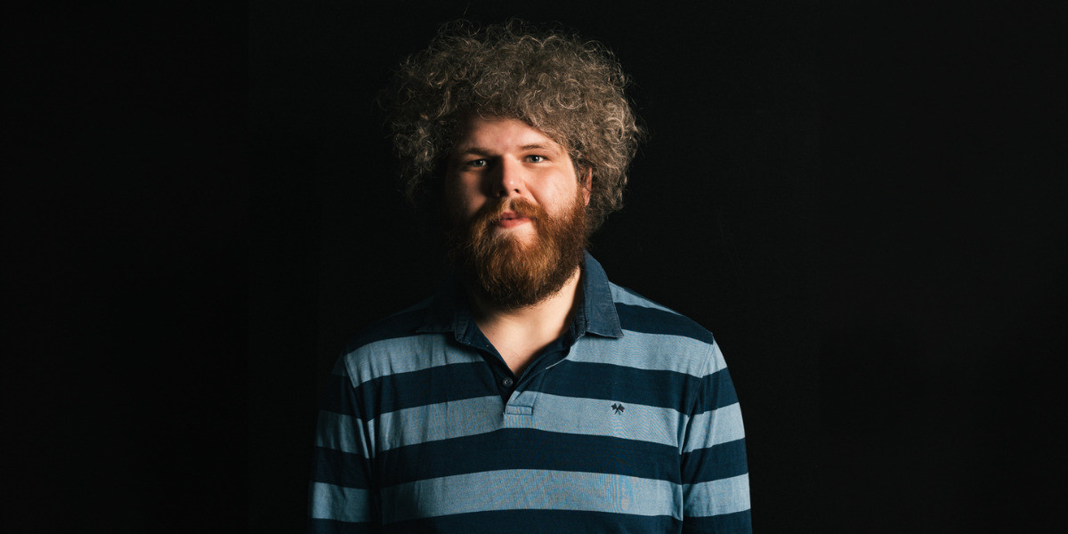 Dave has curly hair and a beard. He is wearing a blue stripy shirt. Dave is looking directly at the camera with a slight smile. The background is black.