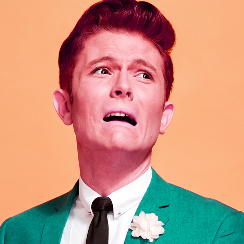 Rhys Nicholson: This is my new material. No Refunds. - A Performer with cherry red hair wearing a black tie and a green suit jacket with a white flower in the button hole looks distressed with a downturned mouth and furrowed brow.
