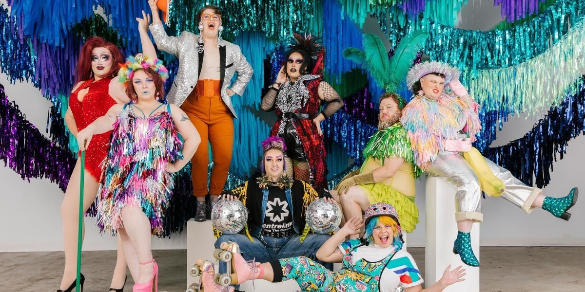 A group of artists pose together dressed in colourful sparkly clothing in front of a streamer installation of shimmery greens and blues. Artists include from left to right Cleo Taurus, Diana Divine, Arthur Nicely, Scott Balls, Say Gah, Calamity Tash, Mathew Barker and Annie Schofield