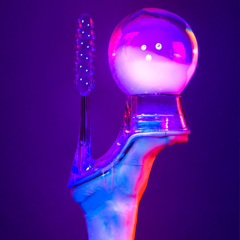 A purple background with a foot wearing a clear plastic platform stiletto is upside down in the centre of the image. There is a large bubble resting on the base of the shoe and six (6) small bubbles stacked on top of each other balancing on the heel of the shoe.