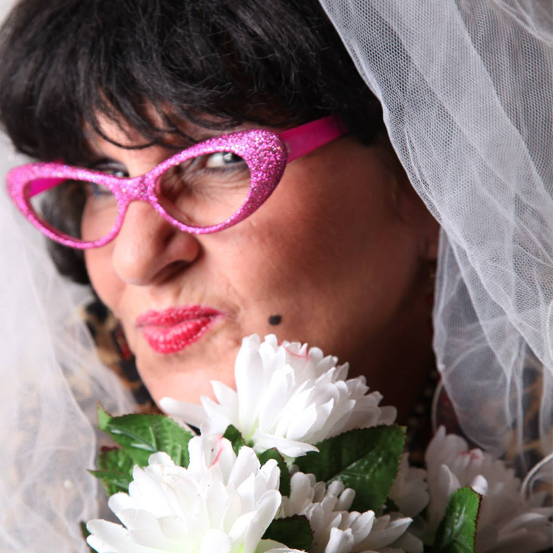 A close up headshot of a woman wearing a white veil and pink glittery framed glasses. She is pouting her lips and is holding a bunch of white flowers.