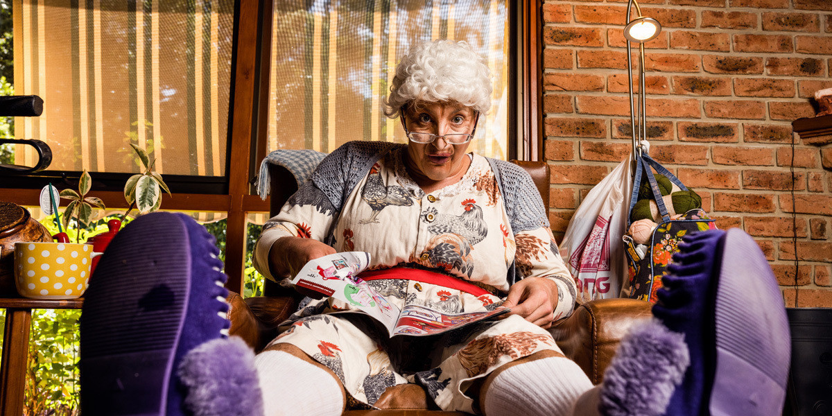 A performer dressd as a granny is sitting on a couch reading a magazine