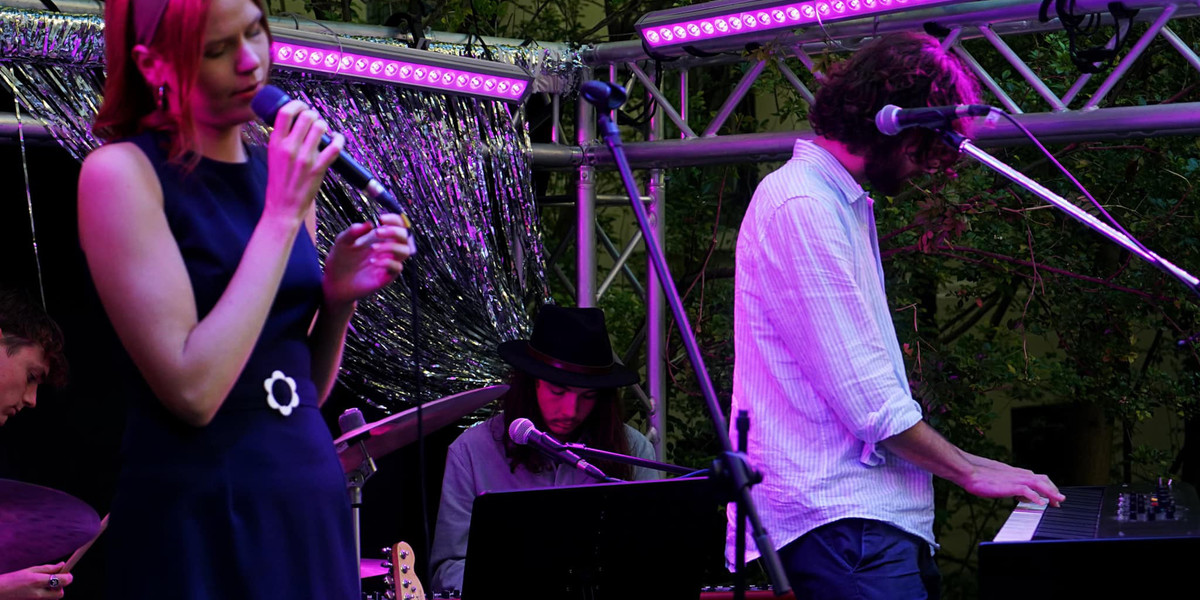 A group of musicians play are playing music on an outdoor stage with soft pink lighting. From left to right they are playing drums, vocals, keyboard, and synthesiser. There is a guitar sitting on a stand behind the synthesiser player who is standing up.