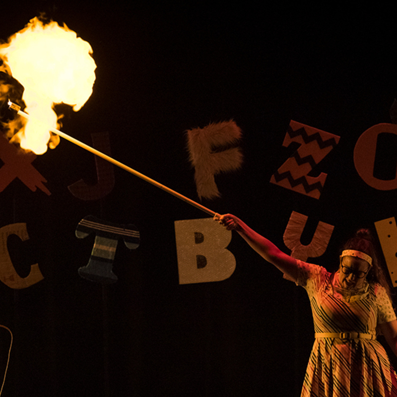 A woman in a striped dress holds a long stick with a candle on the end — she has just exploded a balloon, which has burst into flames.