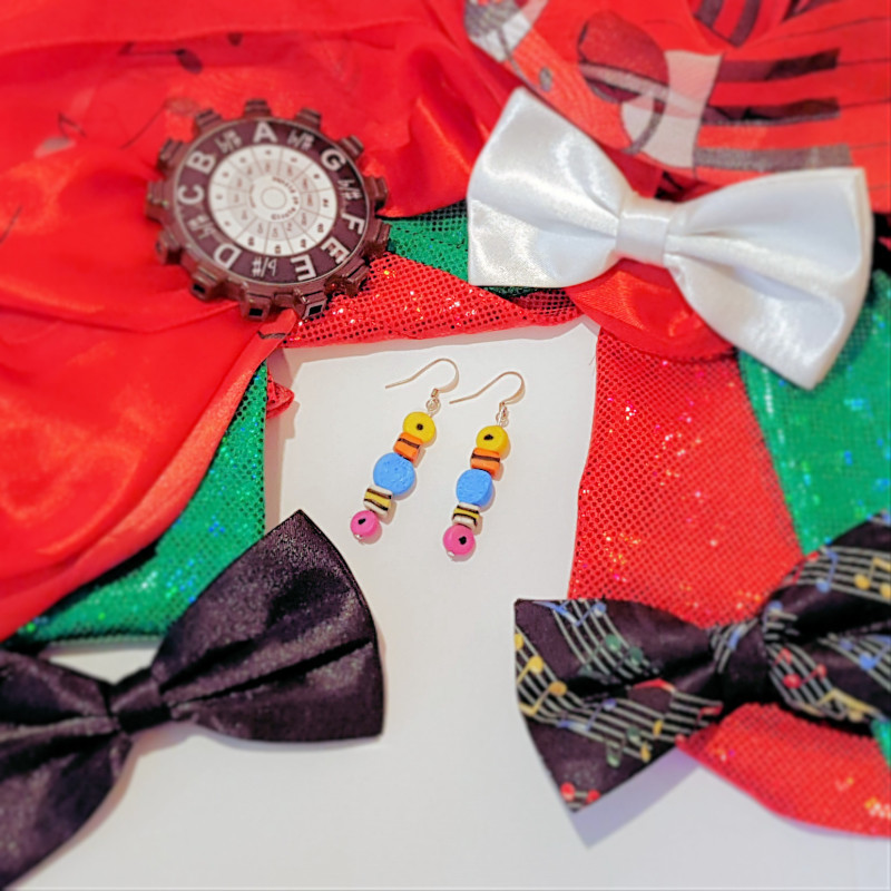 A backdrop of sparkly red and green fabric, three bow ties are spaced around, in the centre is a pair of earrings that look like liquorice allsorts, the top left hand corner is a dark red poker chip.