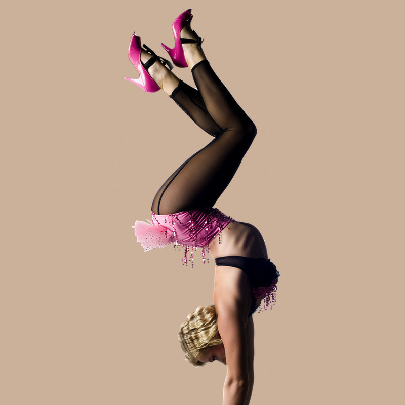 Woman doing a handstand wearing pink high heel shoes and a pink and black costume on a light brown background.