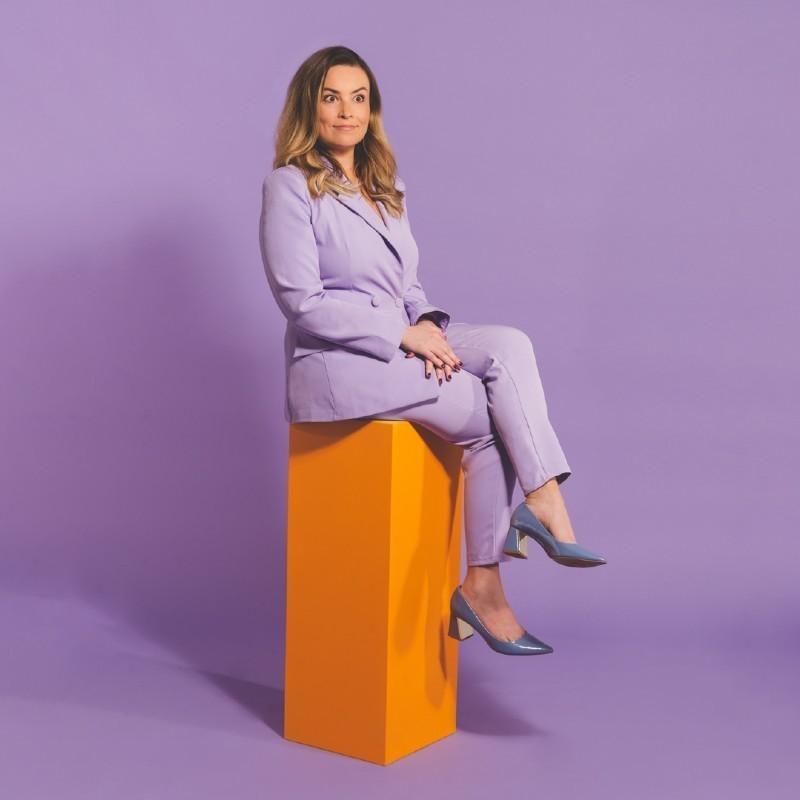 A brown-haired woman wearing a lavender power suit and matching shoes sits atop a bright orange plinth. Although she appears to be poised and smiling like the cover of a self-help book, her eyes are slightly panicked.